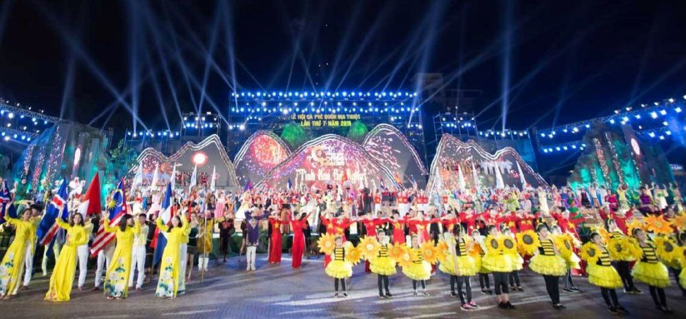 Màn trình diễn sân khấu ngoài trời với ánh sáng rực rỡ, nhiều người mặc trang phục màu sắc và cầm cờ.