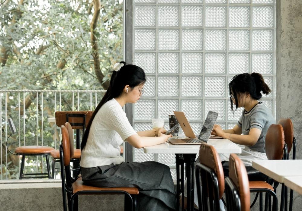 Khám Phá Top 10 Quán Cafe Láng Hạ: View Đẹp, Menu Hấp Dẫn, Giá Cực Hợp Lý 5 Hai người ngồi trong quán cà phê, làm việc trên máy tính xách tay, với cây xanh bên ngoài cửa sổ.