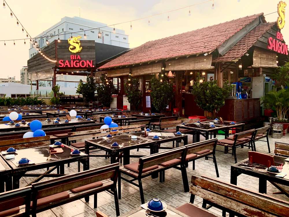 Khám Phá Top 10 Quán Rooftop Quận 1: View Đẹp Ngắm Thành Phố, Trải Nghiệm Thư Giãn Tuyệt Đỉnh 7 Không gian nhà hàng ngoài trời với bàn ghế gỗ, trang trí bóng bay xanh trắng, biển hiệu và đèn treo.