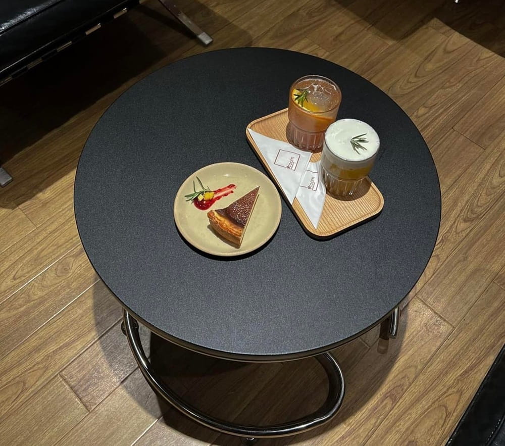 15 Quán Cafe Chùa Láng Với Không Gian Thư Giãn Tuyệt Đẹp 7 Bàn tròn đen với một đĩa bánh ngọt và hai ly nước trên khay gỗ, đặt trên sàn gỗ.