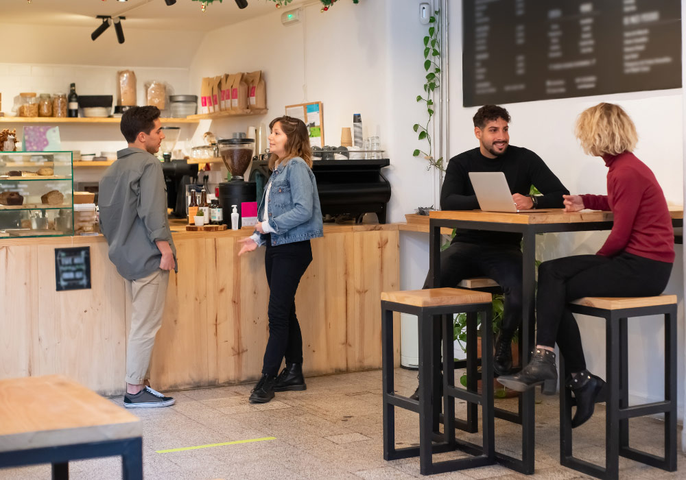 Khám Phá Top 6 Quán Cafe Hot Nhất Tại Thành Thái, Cầu Giấy 15 Quán cà phê với hai người nói chuyện ở quầy và hai người ngồi bàn cao, một người dùng máy tính xách tay.