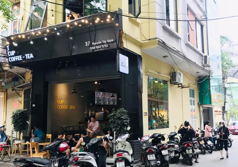 Khám Phá 15 Quán Cafe Hấp Dẫn Trên Đường Nguyễn Thị Định 14 Quán cà phê góc phố với khách ngồi bên trong, nhiều xe máy đậu phía trước, người đi bộ trên vỉa hè.