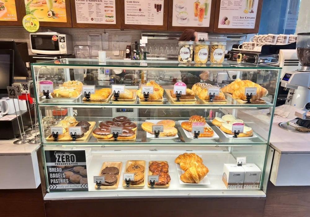 Khám Phá Top Quán Cafe Đẹp Ở Liễu Giai: Đồ Uống Ngon, Không Gian Thư Giãn 6 Tủ kính trưng bày bánh ngọt và bánh mì đa dạng trong một tiệm cà phê, có các món bánh và bảng giá.