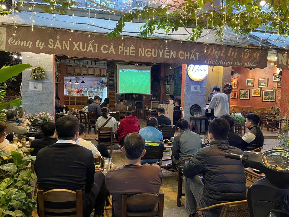 Khám Phá 11 Quán Cafe Đội Cấn Tuyệt Vời Cho Thư Giãn Và Làm Việc 25 Khung cảnh quán cà phê ngoài trời với nhiều người ngồi xem một trận bóng đá trên màn hình lớn.