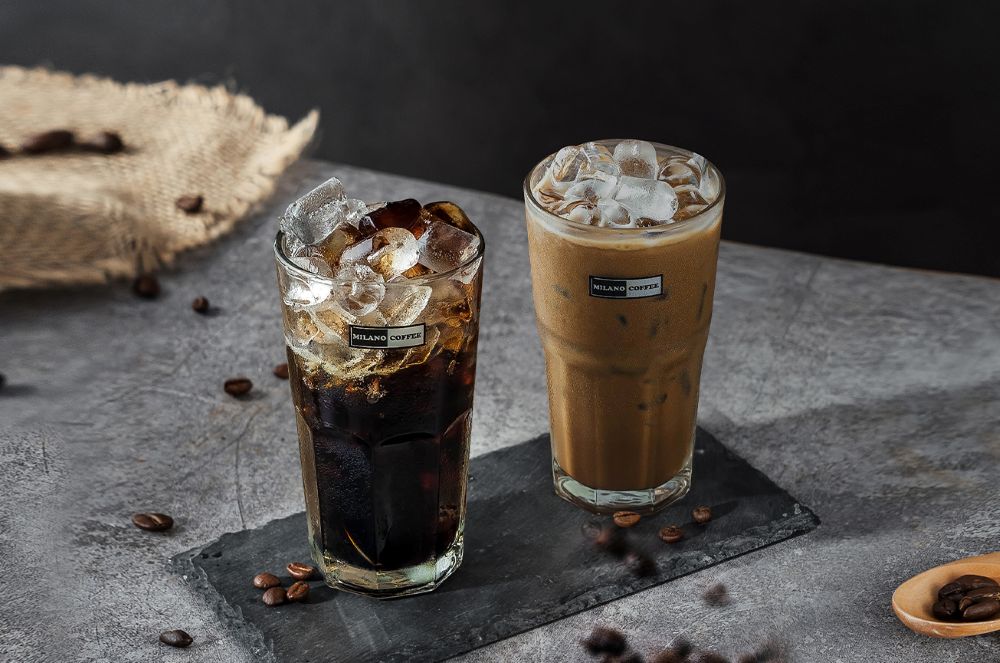 7 Quán Cafe View Đẹp Phạm Văn Đồng Không Thể Bỏ Qua 15 Hai ly cà phê đá trên bàn, một ly đen và một ly nâu, cạnh hạt cà phê và khay gỗ.