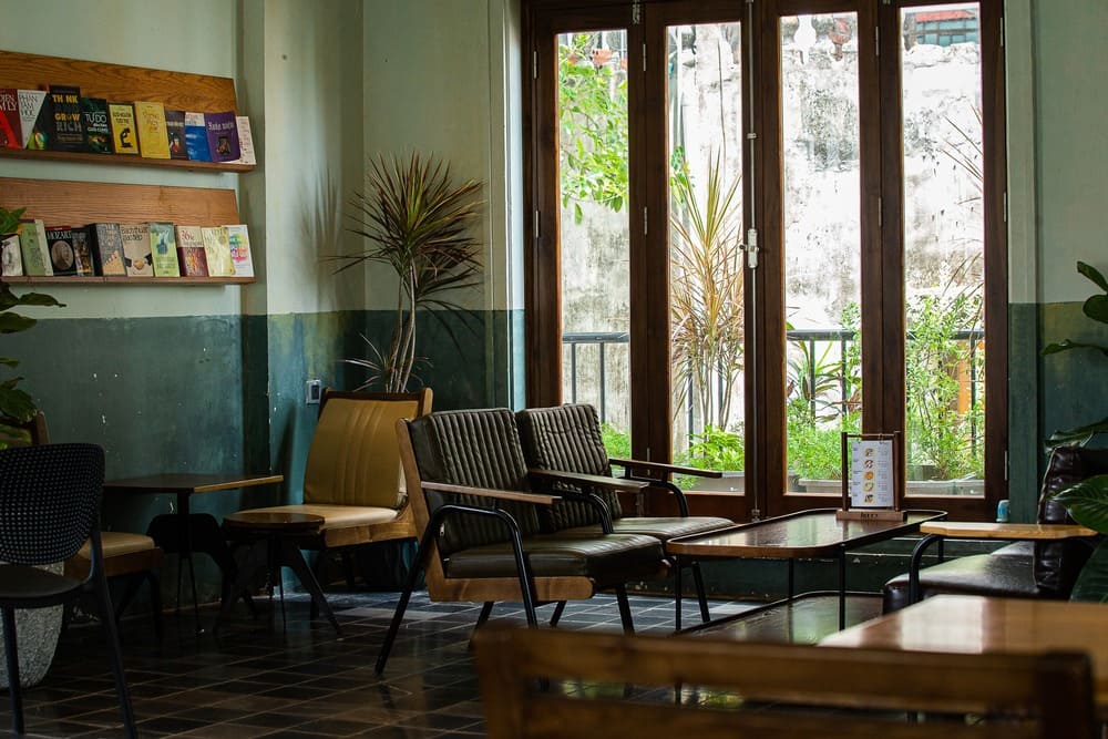 Khám Phá Top 10+ Quán Cafe Quán Thánh Lý Tưởng cho Hẹn Hò, Làm Việc và Thư Giãn 9 Quán cà phê ấm cúng với bàn ghế thoải mái và kệ sách, cửa sổ lớn nhìn ra ngoài cây xanh.