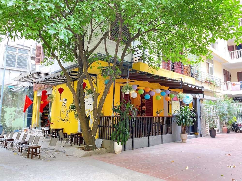 Khám Phá Top 10 Quán Cafe Giảng Võ Hấp Dẫn Nhất Tại Hà Nội 24 Quán cà phê có mặt tiền màu vàng, trang trí đèn lồng và cờ, với cây xanh lớn phía trước và ghế xếp ngoài trời.