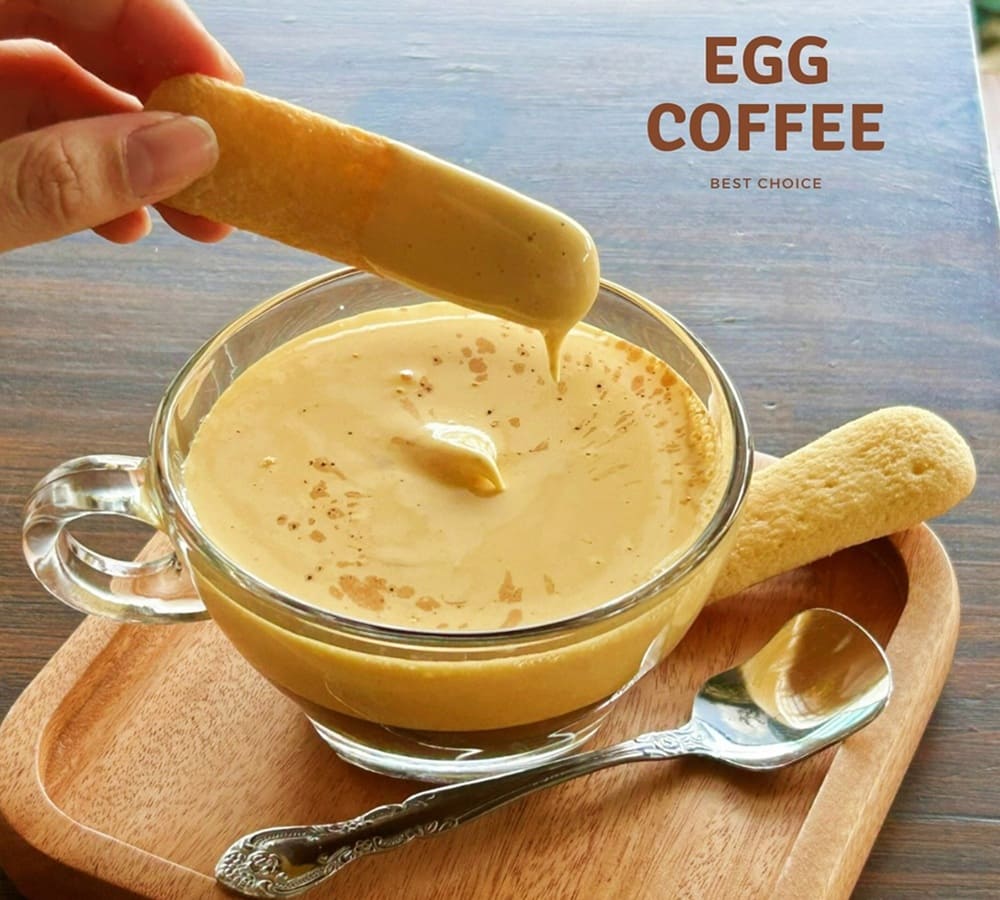 Khám Phá 15+ Quán Cafe View Đẹp, Đồ Uống Ngon Tại Hoàng Cầu 17 Ly cà phê trứng trên bàn gỗ, có người nhúng bánh quy vào, bên cạnh một chiếc thìa bạc.
