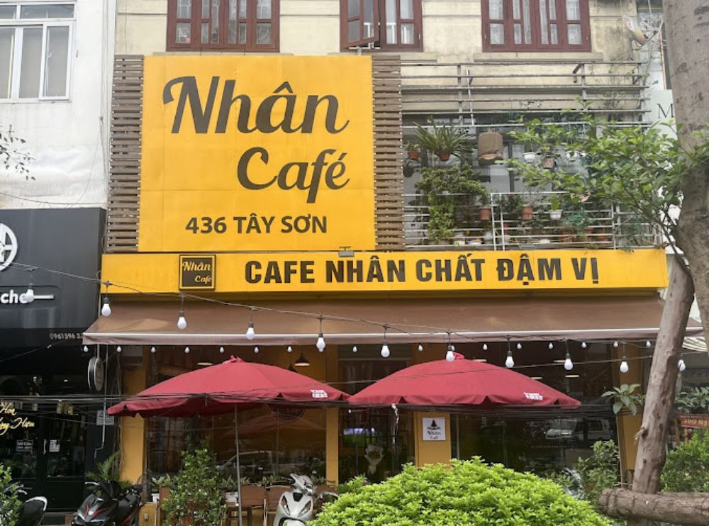 Khám Phá 12+ Quán Cafe Đẹp Ở Tây Sơn Với Đồ Uống Ngon 26 Mặt tiền quán cà phê với biển hiệu màu vàng, cây xanh trang trí và ô dù đỏ phía trước.