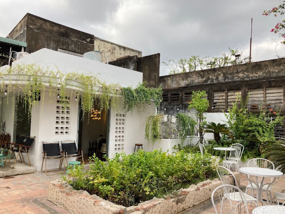 Khám Phá Top 6 Quán Cafe Mở Cửa 24h Tại Quận 3 Cho Người Yêu Thích Cú Đêm 4 Quán cà phê ngoài trời với bàn ghế trắng, cây xanh xung quanh và bức tường cũ kỹ phía sau.
