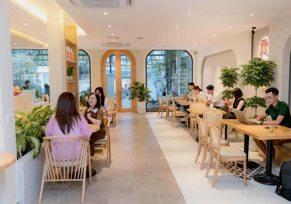 Khám Phá Top Quán Cafe Đẹp Ở Liễu Giai: Đồ Uống Ngon, Không Gian Thư Giãn 5 Quán cà phê với không gian sáng sủa, nhiều cây xanh, khách hàng trò chuyện và làm việc trên máy tính xách tay.
