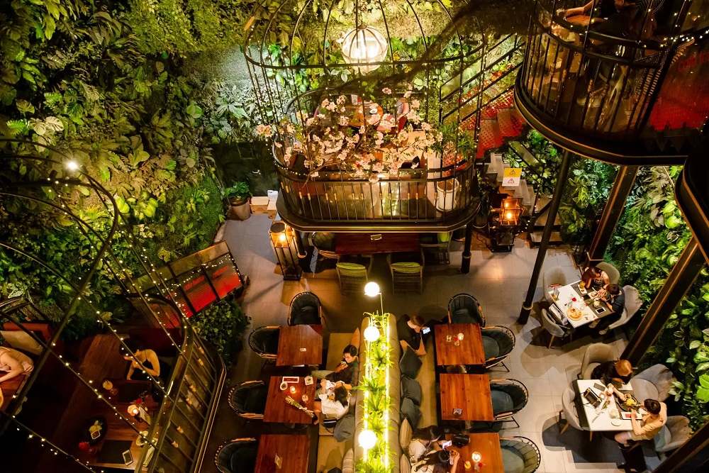 Khám Phá Những Quán Cafe Độc Lạ Thu Hút Giới Trẻ Tại Sài Gòn 26 Không gian nhà hàng với bàn ghế gỗ, cây xanh trang trí và khách ngồi ăn uống, nhìn từ trên cao.