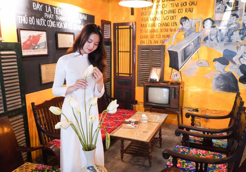 Khám Phá Top Quán Cafe Đẹp Ở Liễu Giai: Đồ Uống Ngon, Không Gian Thư Giãn 4 Người phụ nữ mặc áo dài trắng đứng bên bình hoa sen trong quán cà phê trang trí phong cách retro, có tivi cổ.