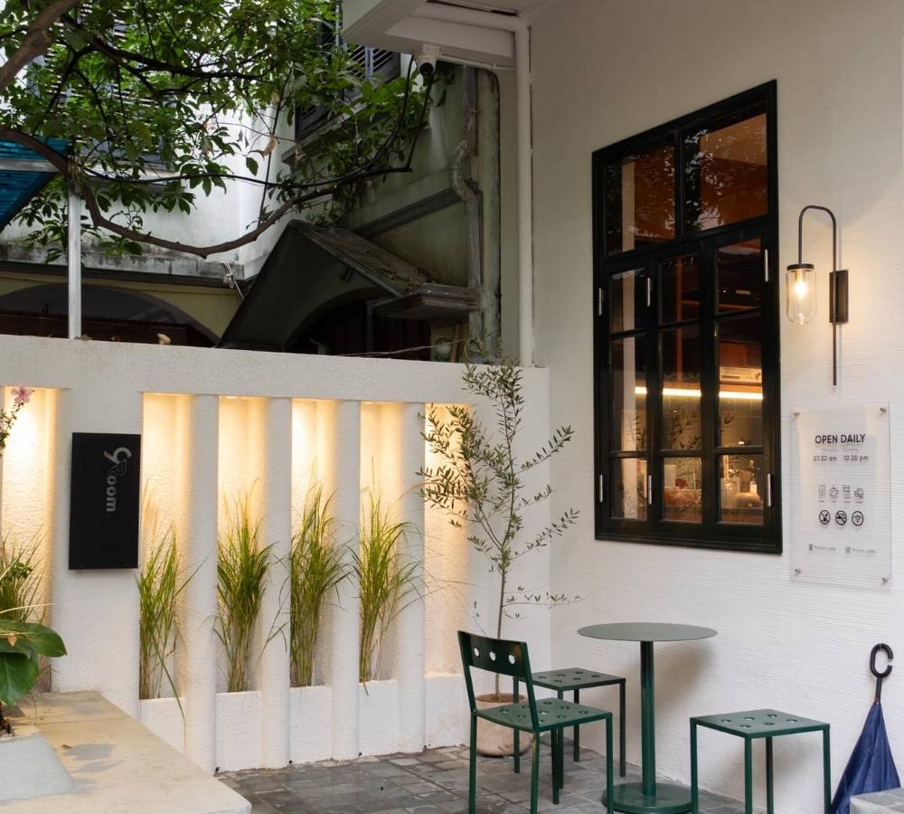 Khám Phá Top Quán Cafe Đẹp Ở Liễu Giai: Đồ Uống Ngon, Không Gian Thư Giãn 18 Quán cà phê với bàn ghế xanh, tường trắng và cửa sổ kính lớn. Cây xanh trang trí trước quán tạo cảm giác thoáng đãng.