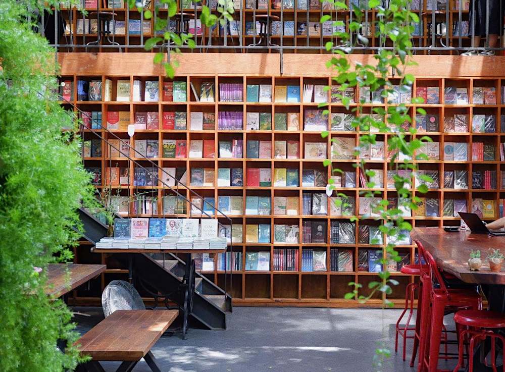 Khám Phá Những Quán Cafe Độc Lạ Thu Hút Giới Trẻ Tại Sài Gòn 11 Kệ sách gỗ nhiều ngăn chứa đầy sách, bên dưới là bàn ghế và cây xanh trang trí trong không gian sáng sủa.