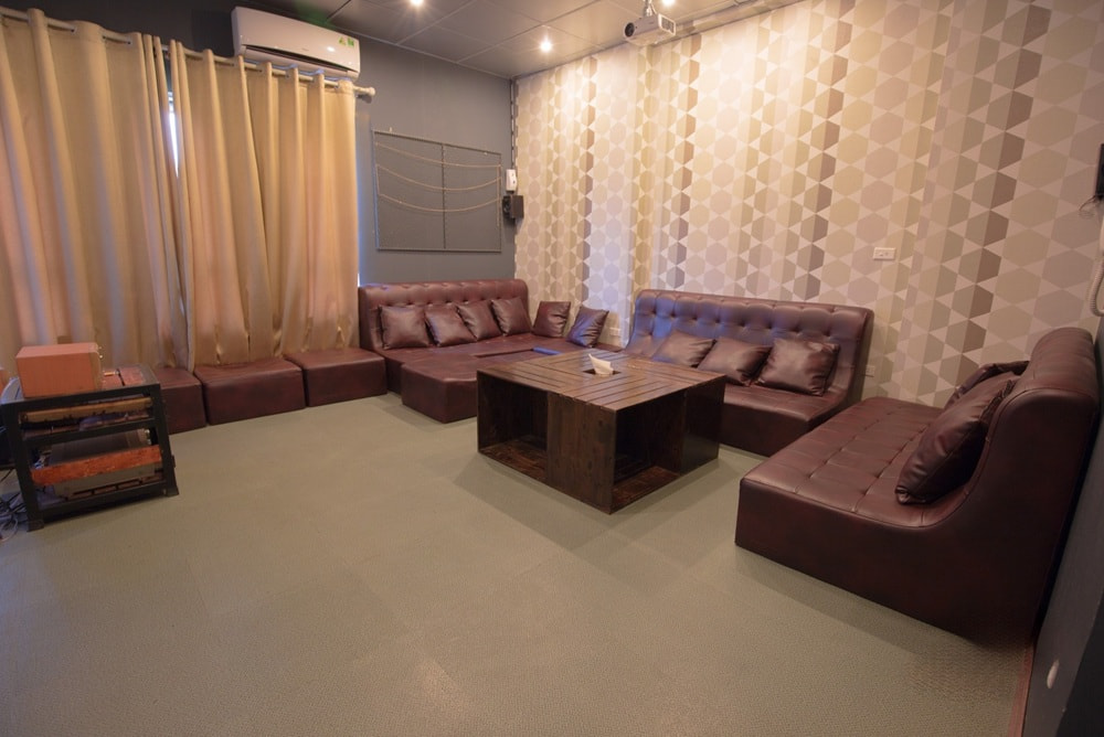 15 Quán Cafe Chùa Láng Với Không Gian Thư Giãn Tuyệt Đẹp 4 Phòng khách với ghế sofa da nâu, bàn gỗ và rèm cửa màu be, tường có họa tiết hình học.