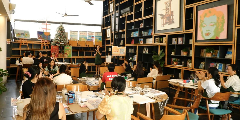 Khám Phá Top Quán Cafe Sách Hot Nhất Quận 1 Bạn Không Thể Bỏ Lỡ Năm Nay 23 Một nhóm người đang tham gia sự kiện trong phòng có giá sách, tranh treo tường và cây thông Giáng sinh.