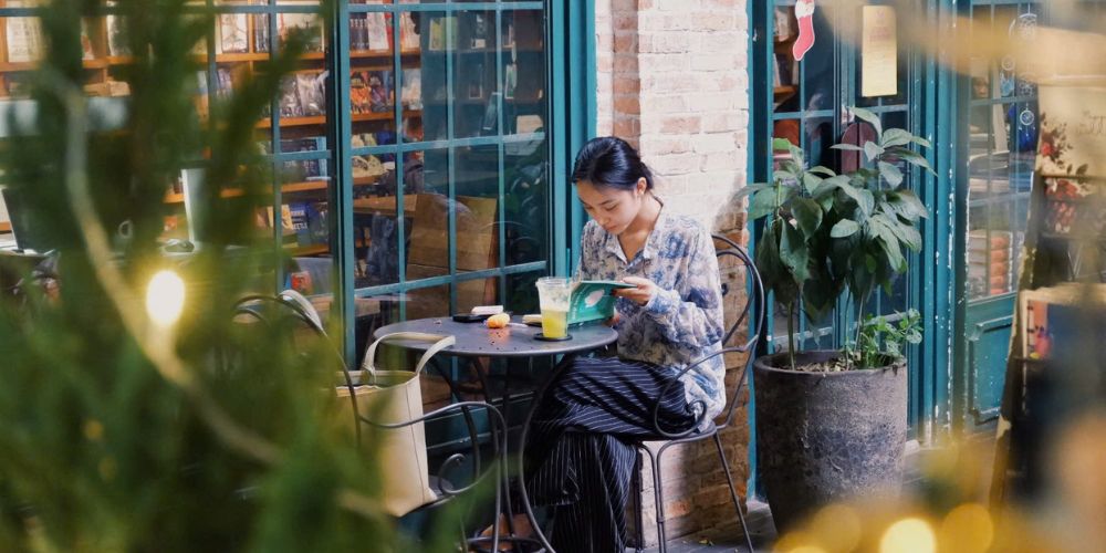 Khám Phá Top Quán Cafe Sách Hot Nhất Quận 1 Bạn Không Thể Bỏ Lỡ Năm Nay 25 Người phụ nữ ngồi đọc sách tại quán cà phê, bên cạnh là cốc nước, xung quanh là cây xanh và kệ sách.