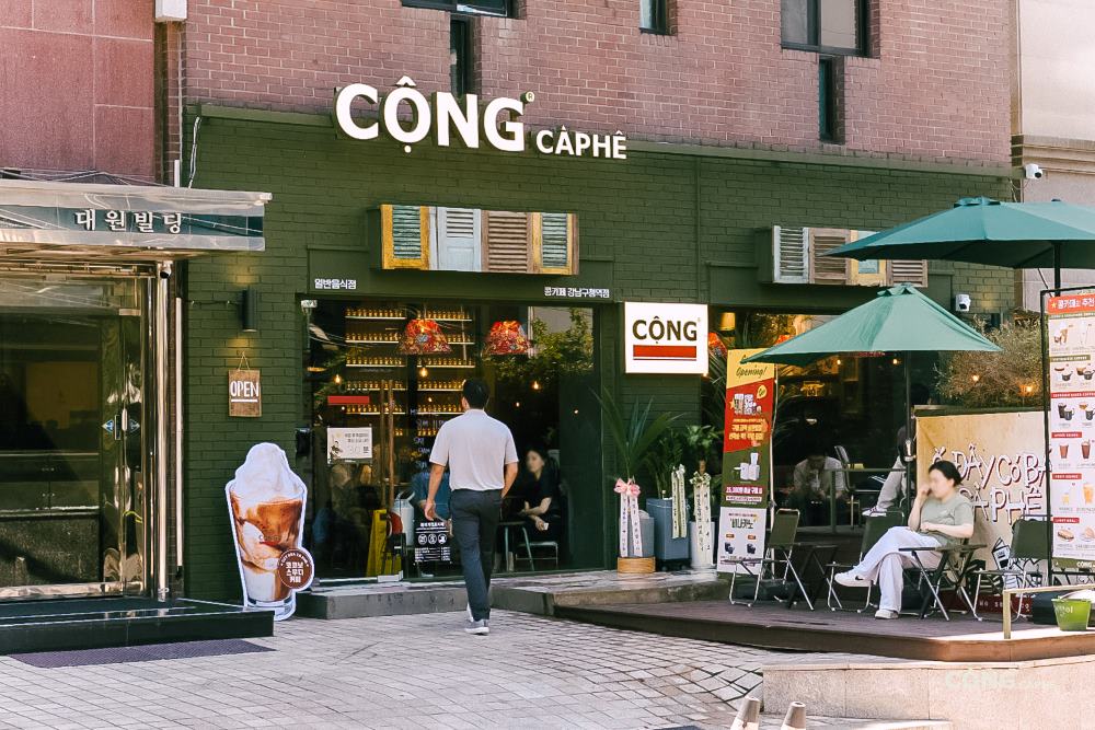 Khám Phá Top 10 Quán Cafe Giảng Võ Hấp Dẫn Nhất Tại Hà Nội 17 Quán cà phê với biển hiệu "Cộng Cà Phê", có khách ngồi ngoài trời dưới dù xanh, người đi bộ phía trước.