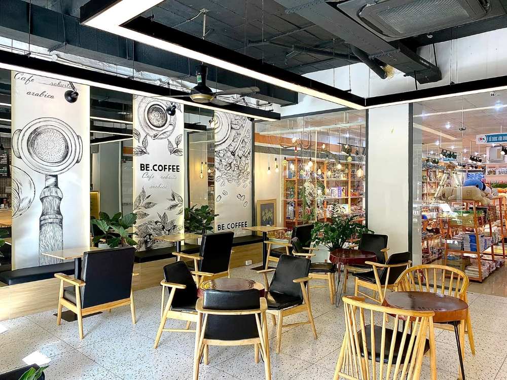 Khám Phá Top 10 Quán Cafe Giảng Võ Hấp Dẫn Nhất Tại Hà Nội 13 Quán cà phê với bàn ghế gỗ, trang trí cây xanh, ánh sáng tự nhiên và cửa kính nhìn ra cửa hàng sách.