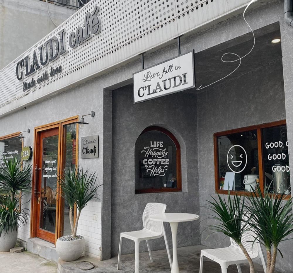 Khám Phá 15+ Quán Cafe View Đẹp, Đồ Uống Ngon Tại Hoàng Cầu 5 Quán cà phê với bảng hiệu "CLAUDI café", có bàn ghế trắng và cây xanh phía trước.