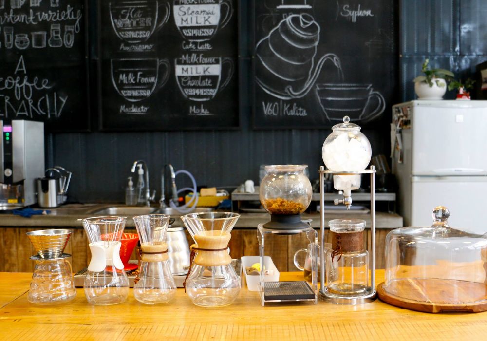 Khám Phá Top 10 Quán Cafe Láng Hạ: View Đẹp, Menu Hấp Dẫn, Giá Cực Hợp Lý 13 Bàn pha chế cà phê với nhiều dụng cụ và bình lọc, nền là bảng đen có vẽ công thức và tủ lạnh trắng.