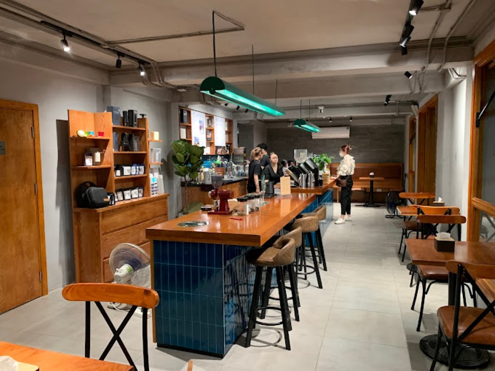 Khám Phá Top 10 Quán Cafe Giảng Võ Hấp Dẫn Nhất Tại Hà Nội 19 Quán cà phê với quầy bar gỗ dài, nhân viên phục vụ, bàn ghế xung quanh, ánh sáng ấm áp và trang trí đơn giản.