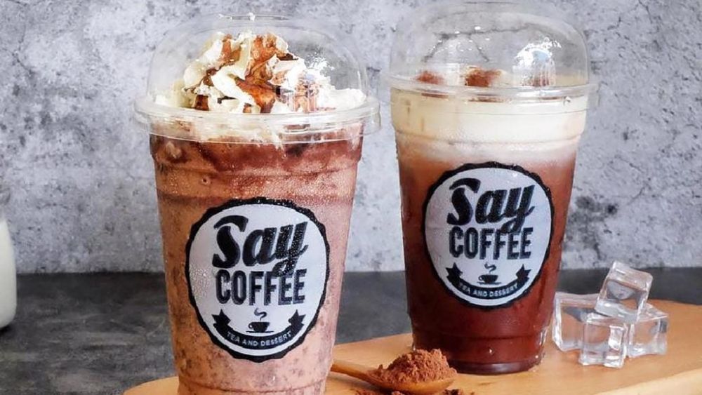 Khám Phá 4 Quán Cafe 24h Ở Bình Thạnh Hoàn Hảo Cho Làm Việc Và Thư Giãn 5 Hai ly đồ uống có nhãn "Say Coffee" trên bàn, một ly có kem, bên cạnh là thìa bột cà phê và đá viên.