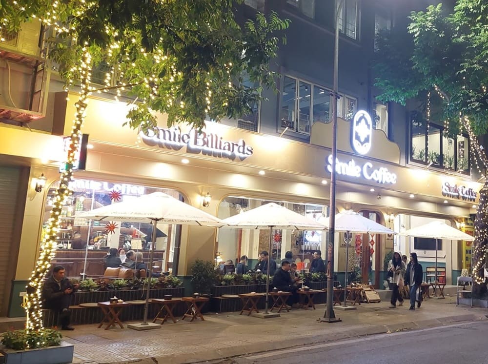 15 Quán Cafe Chùa Láng Với Không Gian Thư Giãn Tuyệt Đẹp 16 Quán cà phê và bida vào buổi tối, với đèn trang trí và khách ngồi dưới ô ngoài trời.