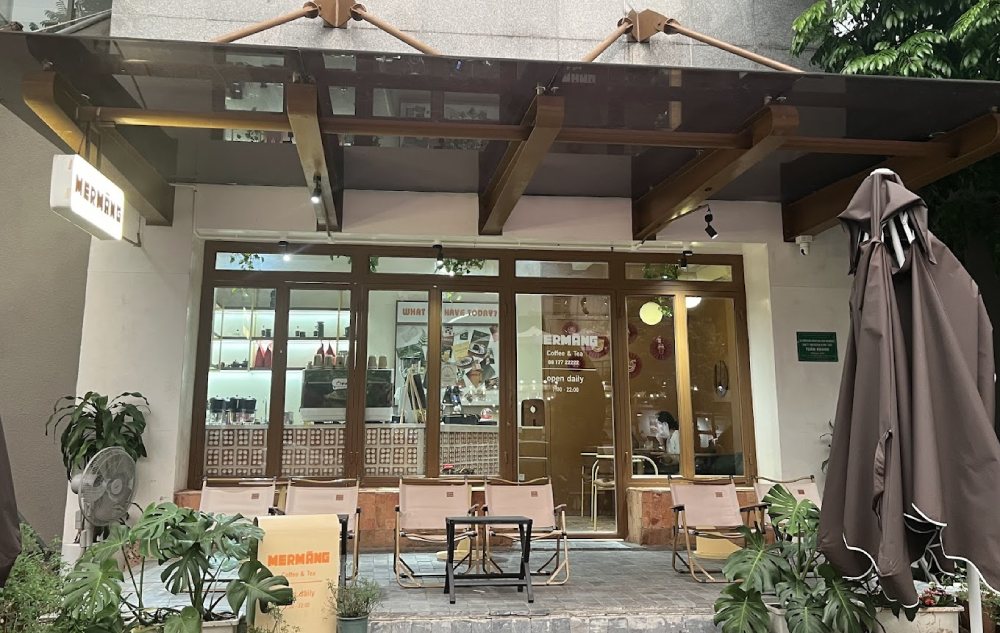 Khám Phá 7 Quán Cafe Tuyệt Đẹp Ở Ngã Tư Sở: View Đẹp, Đồ Uống Ngon, Giá Phải Chăng 18 Mặt tiền quán cà phê với ghế và ô che, cửa kính lớn, cây xanh xung quanh tạo không gian thoáng mát.