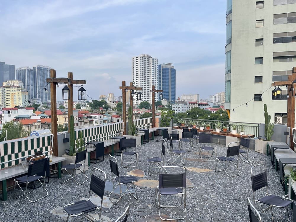 15 Quán Cafe Chùa Láng Với Không Gian Thư Giãn Tuyệt Đẹp 3 Quán cà phê ngoài trời với bàn ghế xếp, cây cảnh và đèn trang trí, nhìn ra nhiều tòa nhà cao tầng.