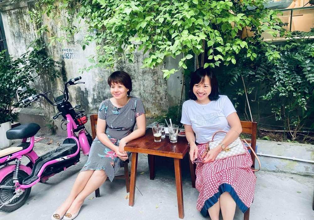 Khám Phá Top Quán Cafe Đẹp Ở Liễu Giai: Đồ Uống Ngon, Không Gian Thư Giãn 12 Hai người ngồi trước bàn nhỏ ngoài trời, bên cạnh chiếc xe đạp điện màu hồng, dưới tán cây xanh.