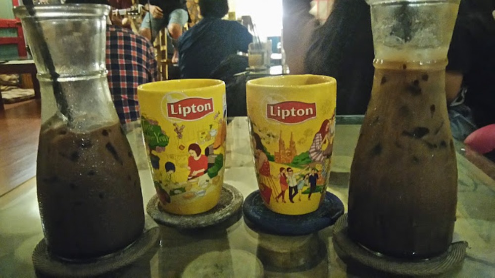 Khám Phá Top 6 Quán Cafe Mở Cửa 24h Tại Quận 3 Cho Người Yêu Thích Cú Đêm 8 Hai ly nhựa màu vàng và hai chai thủy tinh đựng thức uống trên bàn, nền là không gian quán cà phê.