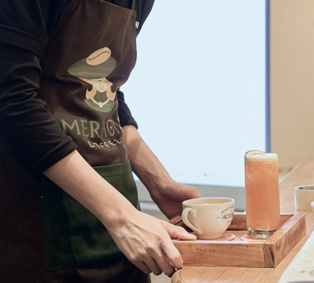 15 Quán Cafe Chùa Láng Với Không Gian Thư Giãn Tuyệt Đẹp 24 Người mặc tạp dề phục vụ khay với một ly cà phê và một ly nước ép trên quầy gỗ.