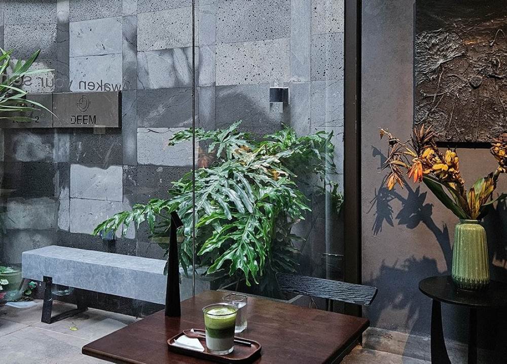 Khám Phá Top 10 Quán Cafe Giảng Võ Hấp Dẫn Nhất Tại Hà Nội 16 Quán cà phê với bàn gỗ, ly trà xanh, chậu cây xanh, tường đá xám và bình hoa trang trí trong không gian hiện đại.