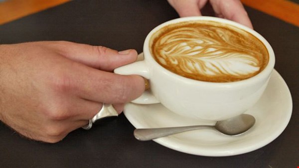 7 Người cầm tách cappuccino với lớp bọt sữa tạo hình nghệ thuật, đặt trên đĩa kèm muỗng nhỏ.