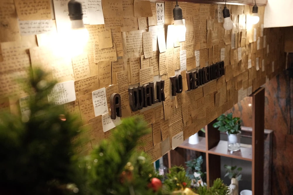 15 Quán Cafe Chùa Láng Với Không Gian Thư Giãn Tuyệt Đẹp 11 Bức tường dán đầy giấy ghi chú với dòng chữ lớn "A Walk to Remember", xung quanh có cây xanh và đèn chiếu sáng.