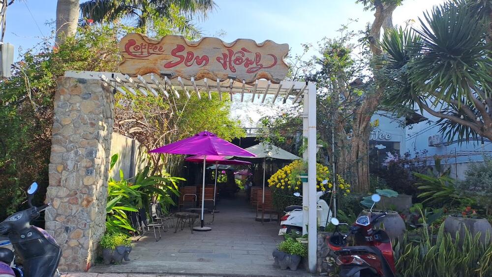 Top 5 Quán Cafe Cá Koi Đẹp Không Thể Bỏ Qua Tại Quy Nhơn 11 Lối vào quán cà phê với biển hiệu bằng gỗ, có dù màu tím và cây xanh xung quanh, xe máy đỗ phía trước.
