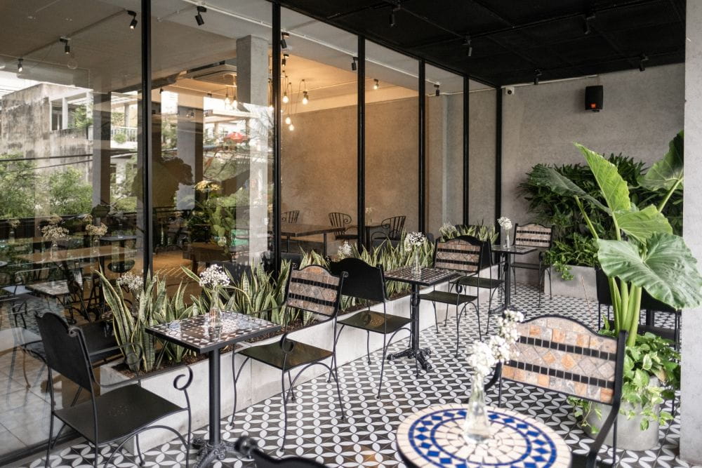 Khám Phá 4 Quán Cafe 24h Ở Bình Thạnh Hoàn Hảo Cho Làm Việc Và Thư Giãn 3 Quán cà phê với bàn ghế ngoài trời, cây xanh trang trí và nền gạch hoa văn đen trắng.