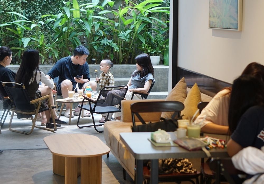 Khám Phá Top Quán Cafe Đẹp Ở Liễu Giai: Đồ Uống Ngon, Không Gian Thư Giãn 22 Nhóm người ngồi trò chuyện trong quán cà phê, xung quanh có nhiều cây xanh, bàn có đồ uống và đồ ăn nhẹ.