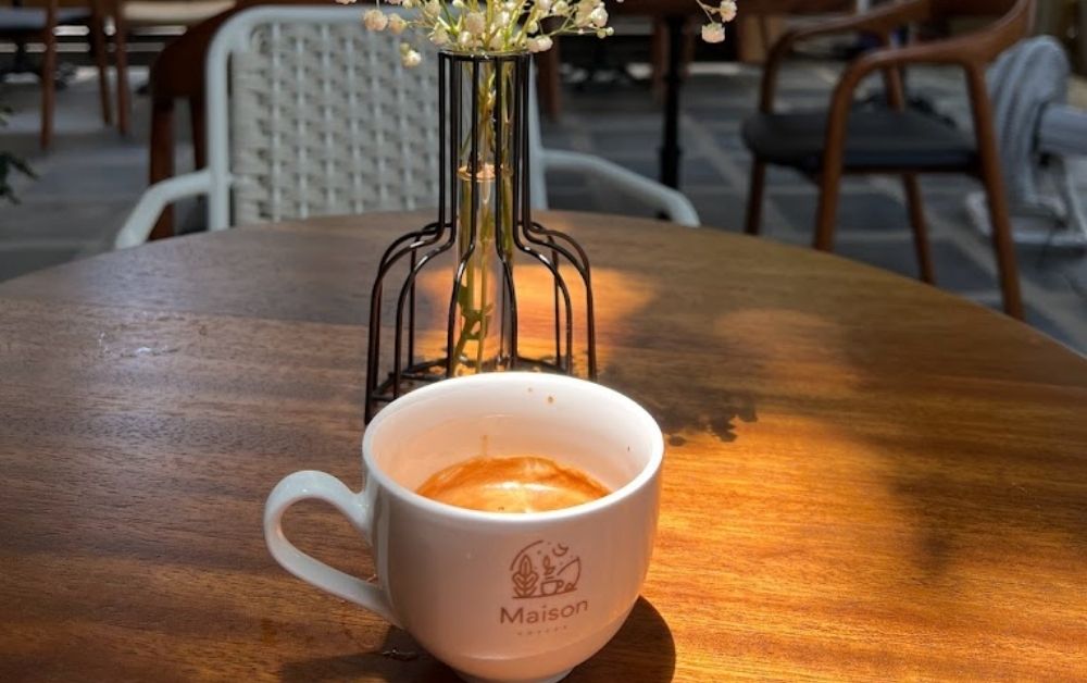 7 Quán Cafe View Đẹp Phạm Văn Đồng Không Thể Bỏ Qua 10 Tách cà phê đặt trên bàn gỗ, phía sau là bình hoa nhỏ và ghế ngồi trong không gian quán cà phê.
