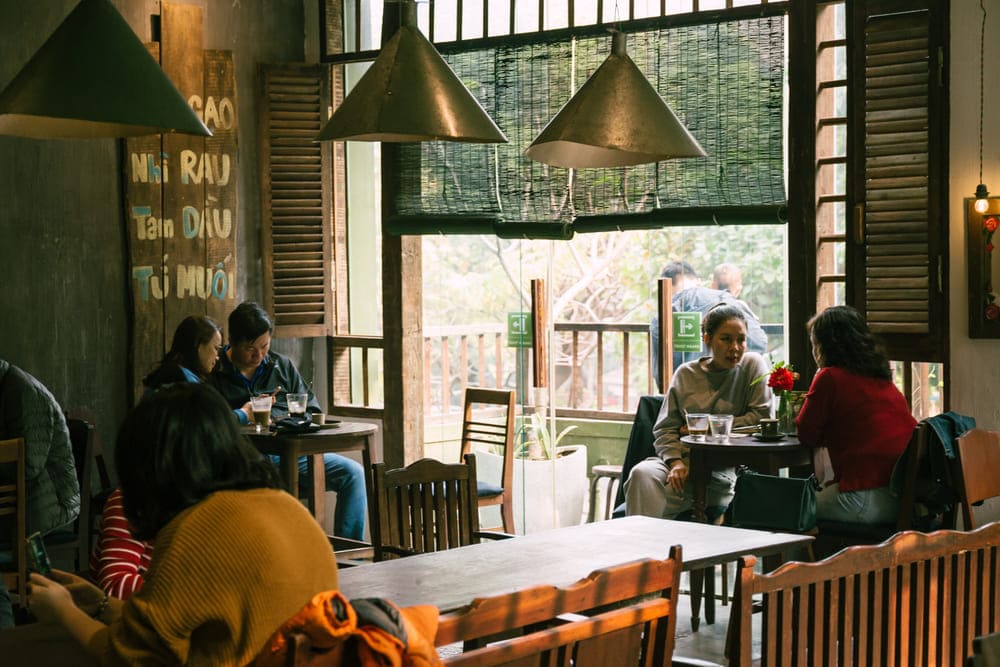 Khám Phá 15 Quán Cafe Hấp Dẫn Trên Đường Nguyễn Thị Định 19 Quán cà phê với nhiều khách ngồi trò chuyện, nội thất ấm cúng và ánh sáng tự nhiên từ cửa sổ lớn.