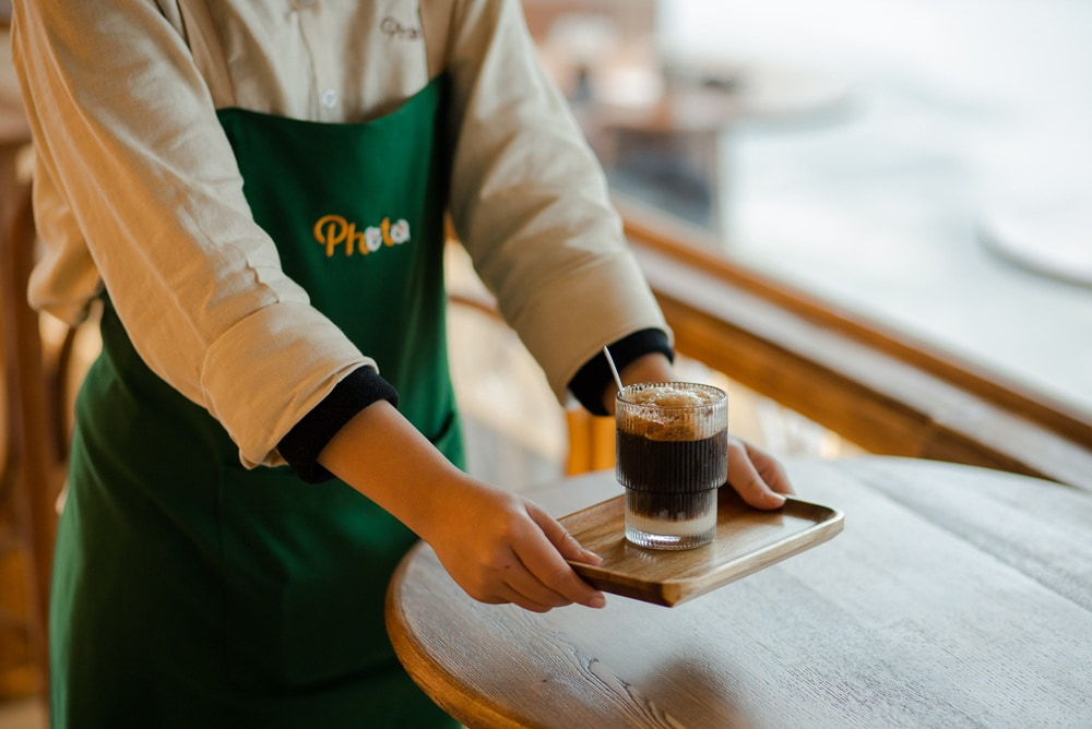 Khám Phá 15+ Quán Cafe View Đẹp, Đồ Uống Ngon Tại Hoàng Cầu 22 Người phục vụ mặc tạp dề xanh lá đang đặt một ly cà phê lên bàn gỗ trong quán cà phê sáng sủa.