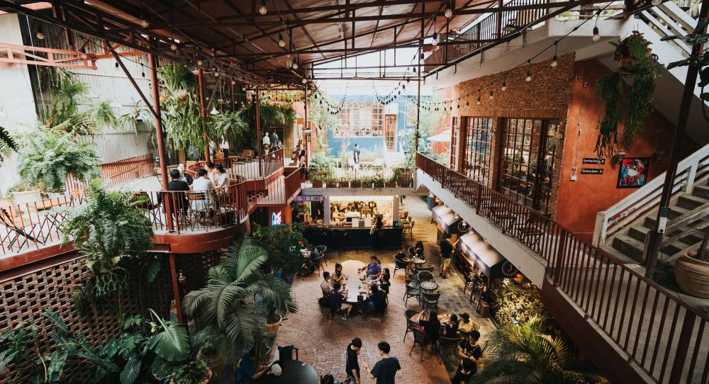 Khám Phá 12+ Quán Cafe Đẹp Ở Tây Sơn Với Đồ Uống Ngon 28 Quán cà phê hai tầng với cây xanh trang trí, bàn ghế ngoài trời, và khách hàng đang trò chuyện.
