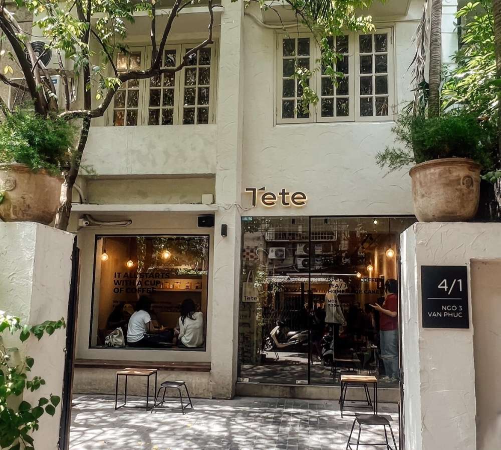 Khám Phá Top Quán Cafe Đẹp Ở Liễu Giai: Đồ Uống Ngon, Không Gian Thư Giãn 15 Mặt tiền của một quán cà phê với cây xanh và cửa kính lớn, có hai người ngồi bên trong.