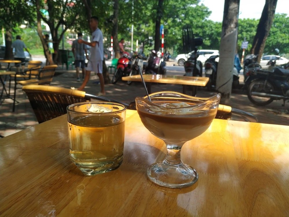 Khám Phá 10+ Quán Cafe Đẹp Ở Đặng Văn Ngữ: Đồ Uống Ngon, Giá Cả Hấp Dẫn 4 Ly trà đá và cốc cà phê sữa đá trên bàn gỗ ngoài trời, với cảnh nền là quán cà phê và cây xanh.