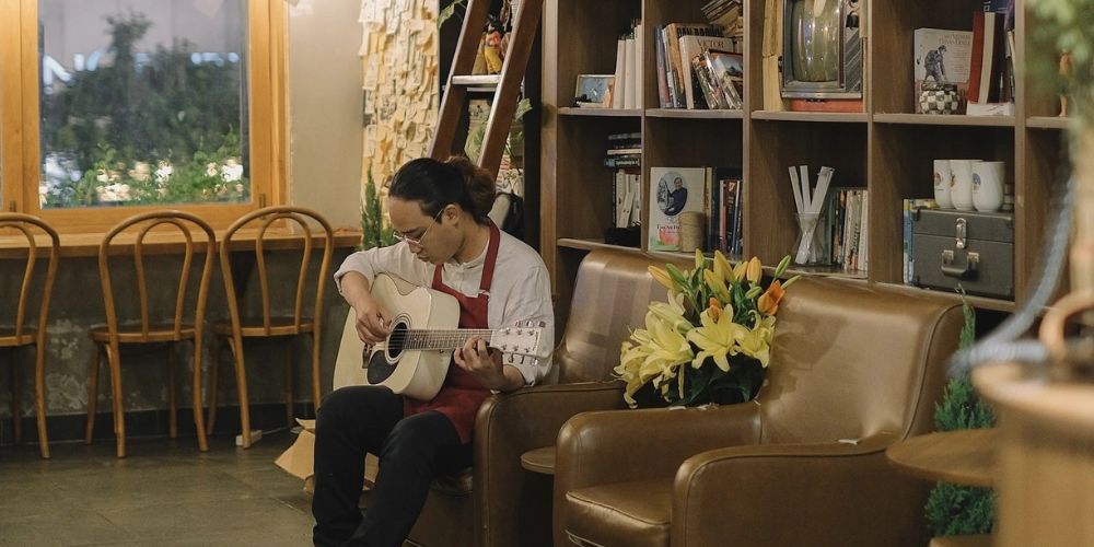 Khám Phá Top Quán Cafe Sách Hot Nhất Quận 1 Bạn Không Thể Bỏ Lỡ Năm Nay 7 Người đàn ông đang chơi guitar trong quán cà phê, ngồi trên ghế sofa bên cạnh kệ sách và bình hoa.