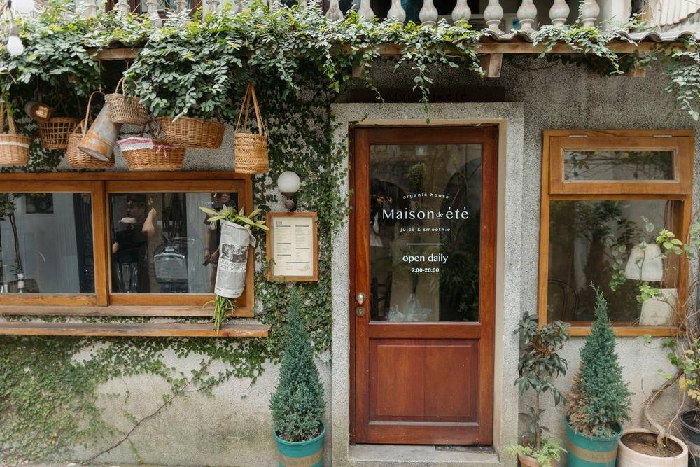 Khám Phá 10+ Quán Cafe Đẹp Ở Đặng Văn Ngữ: Đồ Uống Ngon, Giá Cả Hấp Dẫn 19 Quán cà phê với cửa gỗ, giỏ treo, cây xanh trang trí, không gian ấm cúng và biển hiệu "Maison de été".