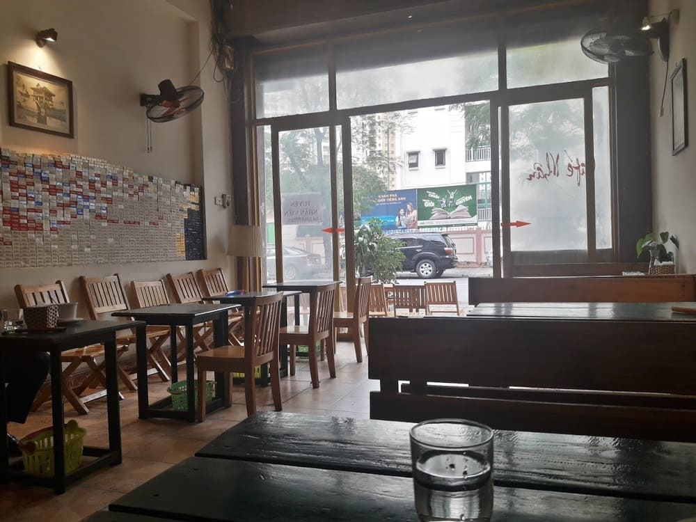 Khám Phá 15 Quán Cafe Hấp Dẫn Trên Đường Nguyễn Thị Định 17 Quán cà phê với bàn ghế gỗ, cửa kính lớn nhìn ra ngoài đường phố, tường trang trí bằng các mảnh ghép màu sắc.