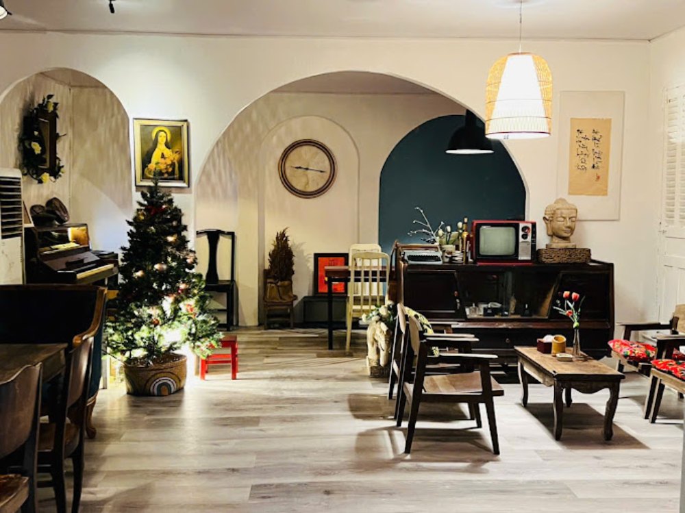 Khám Phá Top 6 Quán Cafe Mở Cửa 24h Tại Quận 3 Cho Người Yêu Thích Cú Đêm 7 Phòng khách cổ điển với cây thông Noel, đàn piano, TV cũ và trang trí nghệ thuật, ánh sáng ấm áp.